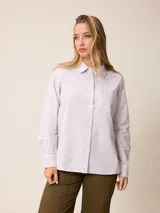 Camisa de manga larga con cuello clásico y cierre frontal de botones. Presenta un diseño de rayas verticales finas en tonos beige y blanco, con un bolsillo frontal en el pecho que incorpora un detalle de parche con rayas horizontales.