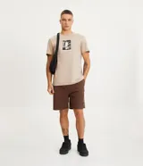 Bermuda masculina color marrón, confeccionada en tejido plano de algodón texturizado con poliéster. Presenta cintura elástica con cordón de ajuste y un tag de marca en el lateral inferior.