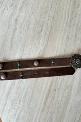 Cinturón de cuero vacuno color marrón, con apliques metálicos circulares y pequeñas piedras turquesas incrustadas. Presenta una hebilla metálica grande con diseño floral grabado.