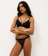 Conjunto de lencería de dos piezas confeccionado en encaje negro con transparencias. El corpiño es de estilo triangular con escote profundo, tirantes dobles y detalles de herrajes dorados. La bombacha es de corte bikini con tiras dobles laterales y adornos metálicos dorados.