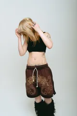 Bermuda de felpa color marrón oscuro con efecto lavado (washed) y detalles en negro en los laterales. Presenta un gran estampado serigrafiado en dorado con tipografía oriental y motivos ornamentales. Tiene cordón de ajuste en la cintura y remaches metálicos cromados en los laterales.