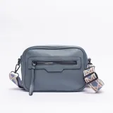 Bandolera de cuero sintético color celeste, con textura granulada, cierre frontal con tirador largo y correa ajustable con diseño geométrico en tonos beige y azul.