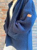 Campera de corderito color verde oscuro, con diseño holgado, cuello alto y manga larga. Presenta una etiqueta de cuero sintético en la manga.