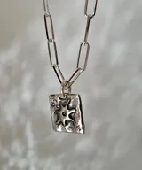 Collar de plata con cadena de eslabones alargados y dije de medalla rectangular con textura rústica, grabado en relieve con forma de estrella y una piedra brillante incrustada en el centro.