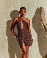 Vestido corto de crochet color marrón con acabado brillante, diseño ajustado al cuerpo, escote halter con tirantes de cadena metálica dorada y abertura frontal pronunciada.