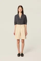 Bermuda de tiro alto en color crudo, confeccionada en lona de algodón. Presenta un corte acampanado con pliegues en la cintura, bolsillos delanteros inclinados y costuras decorativas en contraste color camel.