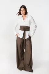 Camisa blanca de algodón con cuello clásico, cierre frontal de botones y puños anchos desmontables. Presenta un diseño con tablas decorativas y terminación redondeada en los laterales.