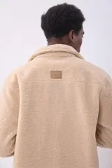 Cazadora de estilo camisa confeccionada en tejido de borreguillo color beige, con cierre frontal de botones, cuello clásico y bolsillo de parche en el pecho.