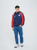 Campera deportiva Umbro del Club Nacional de Football, modelo Venti, para hombre. Diseño en color blanco con mangas azules y detalles de líneas rojas. Cuenta con capucha con cordón ajustable, cierre frontal y bolsillos. Presenta el logo de Umbro y el escudo del club bordados en el pecho.
