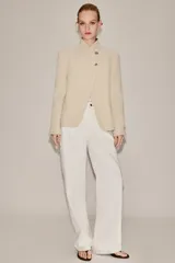 Blazer de corte estructurado en color beige, con cuello mao y cierre frontal cruzado mediante botones. Presenta mangas largas con detalle de botones en los puños y bolsillos de vivo en el frente.