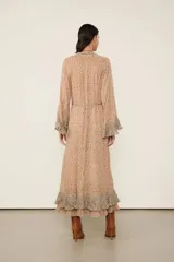 Vestido largo beige estampado con diseño floral, con forro, escote en V con cierre de botones delanteros y mangas largas con puños anchos con volados. Presenta una silueta fluida, detalles plisados y un volante superpuesto en el bajo.