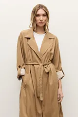Trench de lyocell color beige con solapas amplias, cierre cruzado con botones, bolsillos laterales y mangas largas con presillas en los puños que permiten ajustarlas.