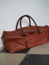 Bolso de viaje tipo duffel bag de cuero color caramelo, con asas de mano y detalles en cuero marrón oscuro. Presenta cierres metálicos y una cremallera exterior con ribete rojo.