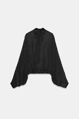 Blusa negra con estampado de lunares blancos, cuello redondo con lazadas y manga larga con puños ajustados. El bajo es ajustable con cordones.