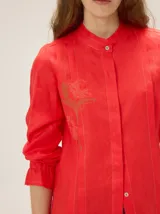 Camisa de lino color crudo, con cuello mao y cierre frontal con botones. Cuenta con detalles de alforzas verticales y diseño bordado floral en el frente, además de mangas 3/4 con volumen y puños elásticos.