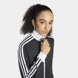 Campera deportiva Adidas Originals, modelo clásico Adicolor Firebird, de color negro con detalles en blanco. Presenta cierre frontal completo, cuello alto tipo embudo, bolsillos laterales con cierre y las icónicas tres tiras blancas en las mangas. El logo del trifolio de Adidas está bordado en el pecho.