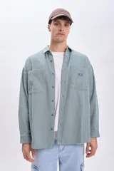 Sobrecamisa verde con rayas verticales blancas, de corte holgado, manga larga, cuello camisero y dos bolsillos frontales con solapa.
