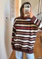 Sweater de punto oversized con diseño de rayas horizontales en tonos marrón, blanco y negro, cuello alto y mangas largas con hombros caídos. Cuenta con aberturas laterales en el dobladillo y una etiqueta roja decorativa en la manga.