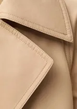 Abrigo tipo trench color beige, de corte oversize y largo hasta la rodilla. Presenta cuello con solapa, cierre frontal cruzado con doble botonadura, cinturón desmontable, dos bolsillos laterales y mangas largas con puños abotonados.