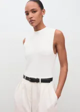 Mono largo de diseño recto y estilo minimalista, color blanco. Presenta cuello perkins, sin mangas con tirantes anchos, detalle de pinzas delanteras y cierre de cremallera en la parte posterior. Incluye bolsillos laterales y un cinturón negro con hebilla metálica.