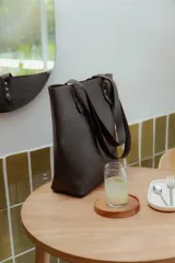 Cartera tipo tote bag de cuero marrón, con diseño minimalista, asas largas y detalles de remaches metálicos en la unión de las asas.