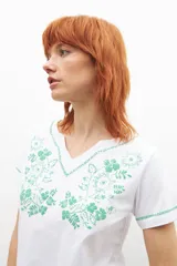 Remera blanca de manga corta con cuello en V, adornada con un bordado floral en color verde menta en el área del pecho y ribetes del mismo color en el cuello y mangas.