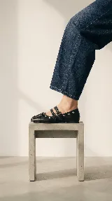 Chatita estilo bailarina de cuero negro, con diseño de doble tira con hebillas metálicas en el empeine y detalle de cordones en la puntera. Presenta ojales metálicos decorativos en los laterales.