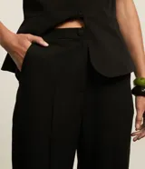 Pantalón de vestir negro, corte carrot (holgado en caderas y ajustado en tobillos), con talle alto, pliegues frontales y cierre tradicional con botón forrado.