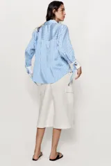 Camisa de cuello solapa y manga larga con rayas verticales celestes y blancas. Cuenta con puños anchos blancos contrastantes, ajustables con hebilla dorada y lazos, y cierre frontal con botones dorados.