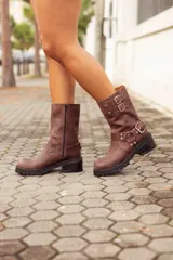 Bota corta de cuero color marrón, con taco cuadrado y punta fina. Presenta una correa con tachas pequeñas y una argolla metálica decorativa alrededor del tobillo. Cuenta con cierre lateral.