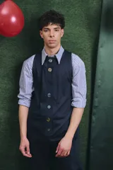 Mono largo de color negro con cierre frontal de botones vintage de distintos colores. Presenta un diseño sin mangas, con bolsillos laterales y una cinta para herramientas en la parte trasera.