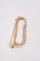 Brazalete multicapa en tono dorado, compuesto por una cadena de eslabones gruesos, una cadena fina tipo serpiente y una cadena delgada con pequeñas perlas blancas intercaladas. Presenta un dije central de nudo metálico y cierre de mosquetón.