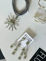 Collar artesanal de la marca Fluss con dije en forma de sol hecho de bronce y alpaca, con incrustaciones de piedra en tono verde y centro de resina con fragmentos. Viene con una tira de gamuza gris.