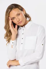 Camisa blanca de manga larga con cuello y tapeta de medio botón. Presenta dos bolsillos frontales con tapa y botones metálicos. Las mangas tienen presillas para sujetar el puño enrollado.