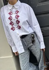 Camisa de algodón blanca con corte clásico, cuello camisero y cierre frontal con botones. Presenta un bordado geométrico en color bordó en la parte delantera y puños abotonados.