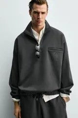 Sudadera de corte relajado color negro, con cuello alto y cierre frontal de cremallera. Presenta un bolsillo de vivo en el pecho con un estampado a contraste y acabados elásticos en puños y ruedo.