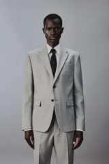 Blazer slim fit de color gris, con cuello de solapas de muesca, cierre frontal de dos botones, bolsillos laterales con solapa y bolsillo de vivo en el pecho. Presenta mangas largas con botones en los puños y aberturas en la espalda.
