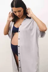 Vestido camisero maternal de color gris claro, con cuello tipo polo en contraste azul marino, cierre frontal con botones y bordado de un corazón pequeño en el pecho. Presenta mangas cortas y bolsillos laterales.