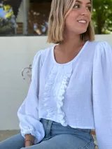 Blusa blanca de algodón con textura arrugada, cuello redondo, mangas largas abullonadas con puños elásticos y cierre frontal con botones y volantes.