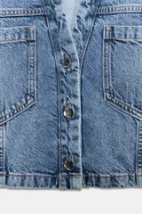 Camisa corta de denim en color azul claro, con cuello solapa, escote en V y manga corta. Presenta bolsillos delanteros de parche con solapa y cierre frontal de botones metálicos.