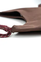 Cartera tipo hobo de gamuza color marrón, con costura central vertical y correa de cuerda trenzada en tonos borró y gris con nudos decorativos en los extremos.