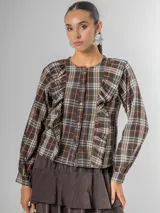 Camisa de manga larga con cuello redondo y cierre frontal de botones. Presenta volados verticales a lo largo del frente y mangas abullonadas. El tejido es de tartán liviano en tonos marrón oscuro, beige, naranja y azul.