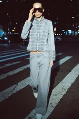 Chaqueta de denim con efecto lavado, corte entallado y cuello redondo. Presenta un diseño de estilo militar con cierre frontal de cremallera metálica y aplicaciones decorativas de botones y costuras en el delantero y los puños.