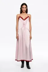 Vestido midi de corte recto y estilo lencero, confeccionado en tejido satinado color rosa. Presenta escote en pico con detalle de encaje en color bordó, tirantes finos, lazo frontal y costuras decorativas verticales a lo largo de la prenda. El ruedo también cuenta con un borde de encaje a tono con el escote.