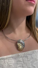 Gargantilla tipo choker rígida de metal plateado con un dije central en forma de gota compuesto por dos piezas texturizadas, una en tono plateado y otra en tono dorado.