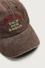 Gorra de seis paneles marrón con efecto lavado, visera curva y texto bordado en relieve en la parte frontal en tonos rojo y amarillo.