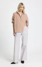 Camisa de corte recto y diseño oversize, con estampado de rayas verticales en tonos marrón y blanco. Presenta cuello clásico estructurado, mangas largas con puños anchos, hombros caídos y bajo asimétrico con abertura lateral. Cierre frontal mediante botones.