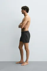 Bañador de hombre color negro, de largo medio, confeccionado en tejido técnico. Presenta cintura elástica ajustable con cordón interior, trabillas laterales, bolsillos frontales y bolsillo trasero con cierre de cremallera.