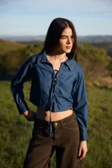 Camisa crop de denim azul con cuello camisero y cierre frontal mediante cordones ajustables a través de ojales. Presenta mangas largas con puños abotonados.