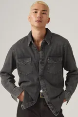 Camisa de jean estilo western en color gris lavado, con cuello clásico, cierre frontal con botones y dos bolsillos delanteros con solapa y botón. Presenta etiqueta roja distintiva en el bolsillo izquierdo.